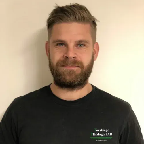 Man med kort, ljusbrunt hår och tjockt skägg står framför en ljus vägg. Han bär en svart t-shirt med företagets logotyp. Ansiktet är vänligt och avslappnat.